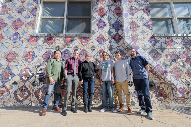 Six individuals stand together in front of a building with pink and purple diamond tiling